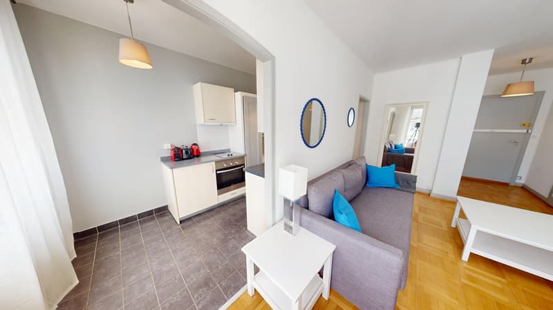 Open plan living room and kitchen, gray couch, white table, gray tile floor, white cabinets, oven, coffee maker, lamps