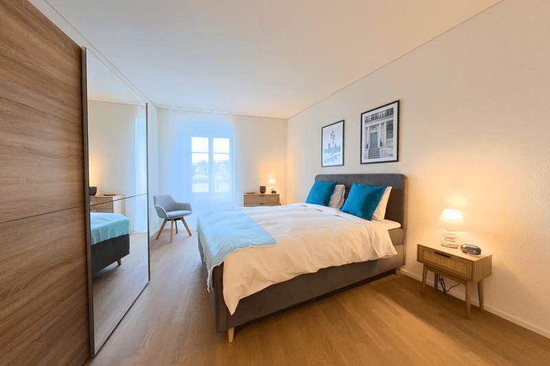 Bedroom: large bed, blue pillows, white linen, nightstand, small lamp, chair, mirror, window with curtains, wooden flooring, two framed pictures on the wall