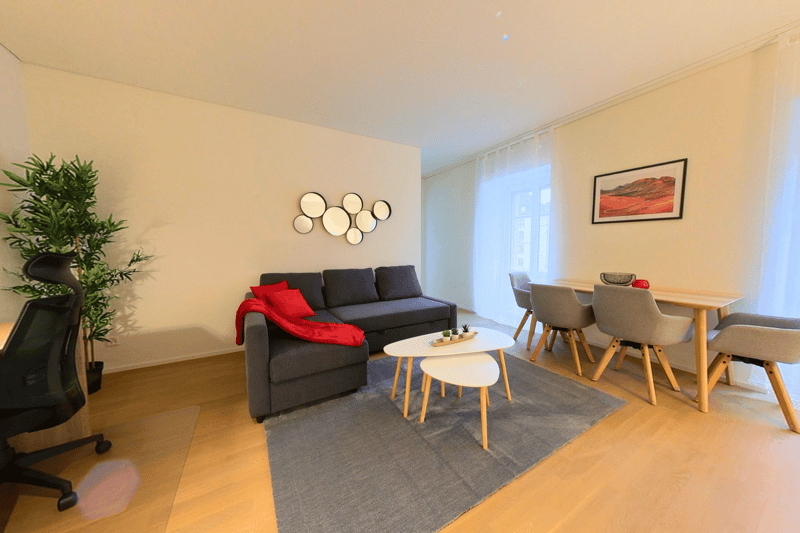 Spacious living room with a black sofa, a red throw blanket, a coffee table with decorative items, a dining table with four chairs, and a framed artwork on the wall. The room also features a potted plant and a window, suggesting the presence of a balcony