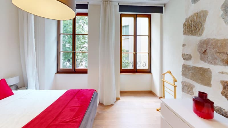 Spacious bedroom with large windows overlooking a balcony, wooden window frames, white walls, red bedding, and a stone accent wall