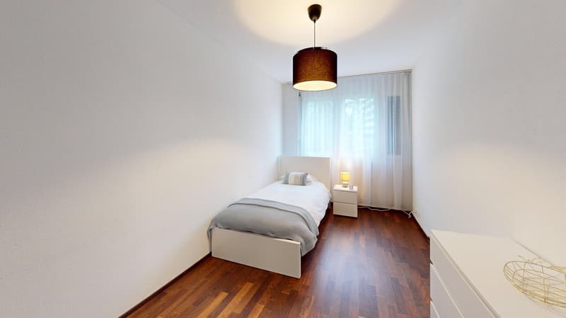 Spacious bedroom with hardwood flooring, white walls, a single bed with gray bedding, a nightstand, and a pendant light fixture with a brown shade.