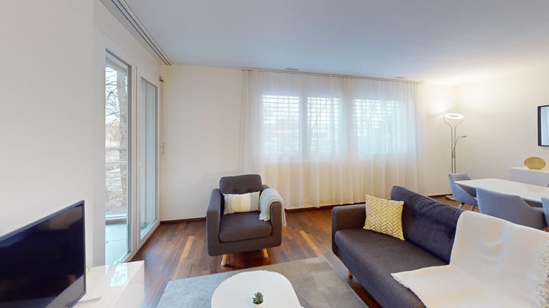 Spacious living room with large windows, hardwood floors, gray sofa and armchair, decorative pillows, and a small coffee table with a plant. The room has access to a balcony or terrace through sliding glass doors.