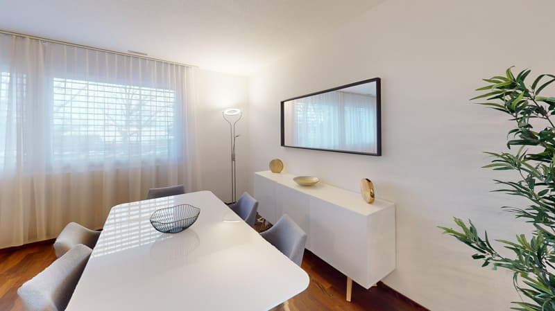Spacious dining area with large windows, white table, gray chairs, wall-mounted mirror, and decorative items on the sideboard.