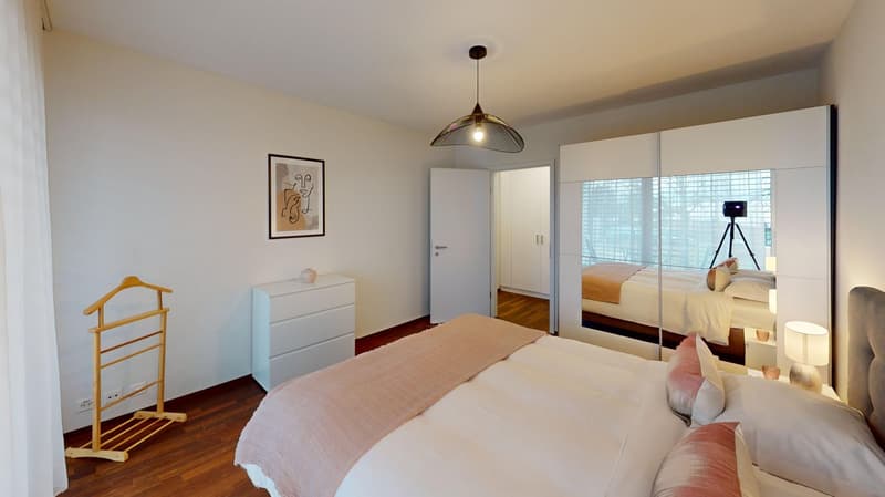 Spacious bedroom with hardwood floors, white walls, large windows, a pendant light fixture, a dresser, and a clothes rack.