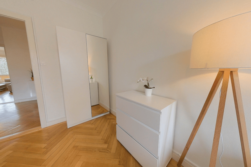 Spacious bedroom with hardwood flooring, white walls, a dresser, and a full-length mirror. The room has a warm and minimalist feel.