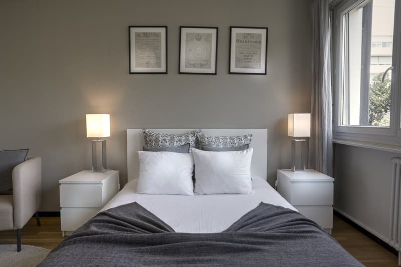 Bedroom with a bed, nightstands, lamps, and a chair. Three framed pictures on the wall. A window with a view of trees outside.