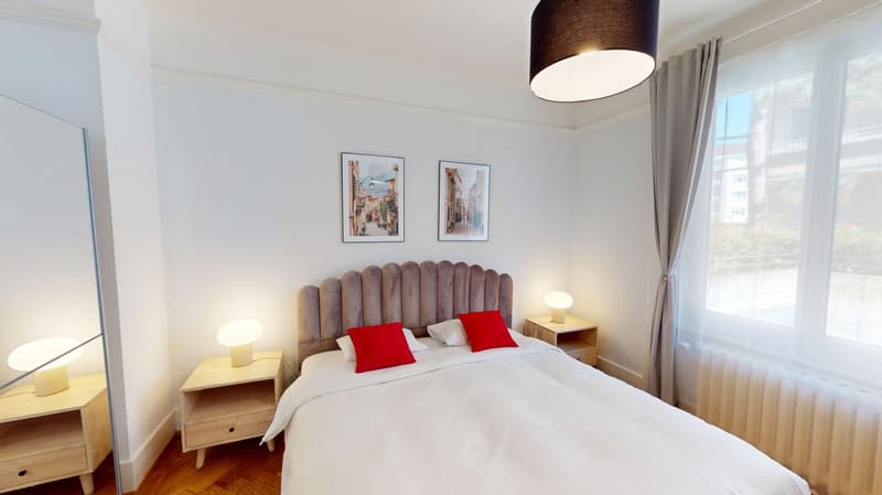 King size bed, white linens, red pillows, lamps, mirror, radiators, windows, photos on wall
