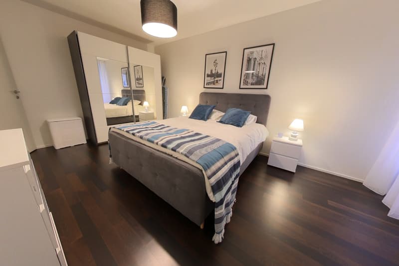 Spacious bedroom with a large bed, dark gray headboard, white nightstands, and framed artwork on the walls. The room has hardwood floors and a pendant light fixture. There is also a wardrobe cabinet visible.