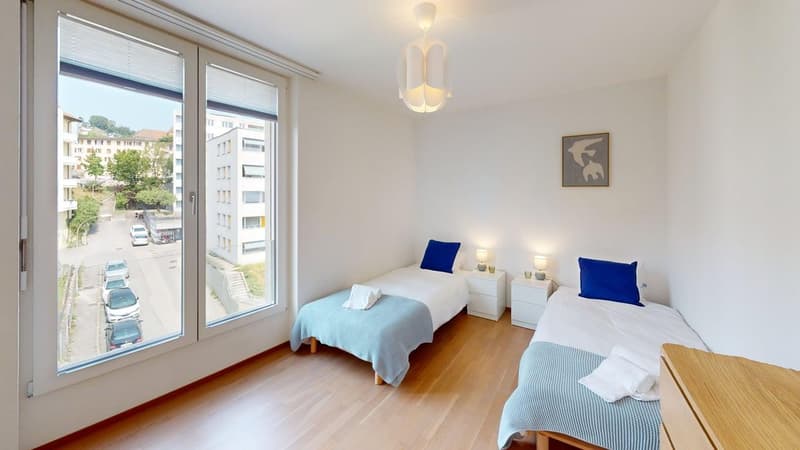 Two beds with white bedding and blue pillows, white nightstands with lamps and cups, large window, wooden floor, framed picture on the wall