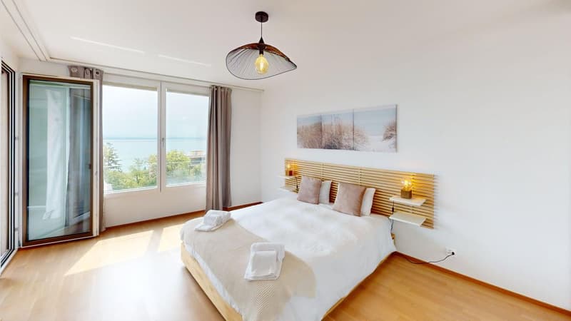 Large bedroom with large windows, sliding glass doors, wooden bed, white linen, wooden floor, nightstand with lamps