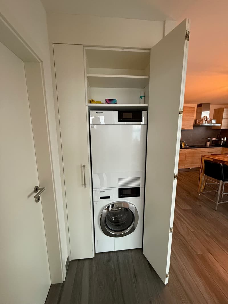 Stacked washer-dryer, wooden flooring, cabinet, and open door leading to the kitchen