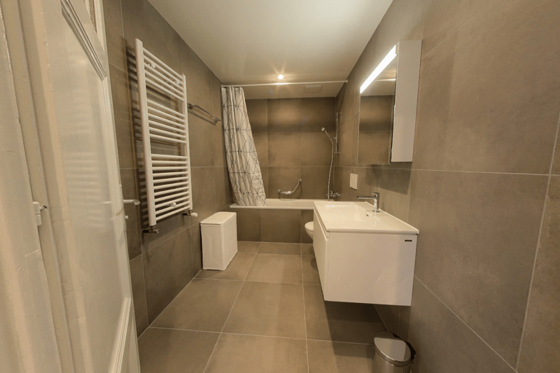 Bathroom: grey tiles, white bathtub, white sink, toilet, towel heater, stainless steel trash can, mirror