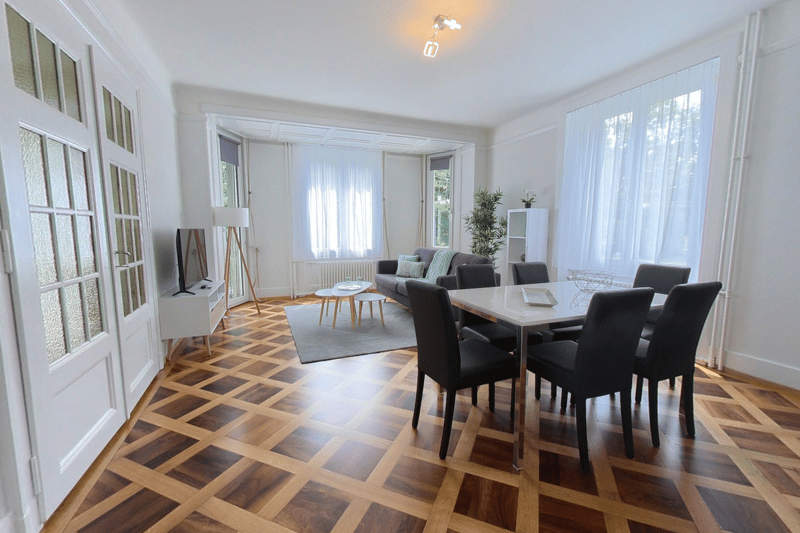 Living room: hardwood flooring, couch, TV, coffee table, dining area with table and chairs, white walls, windows, large radiator, stand
