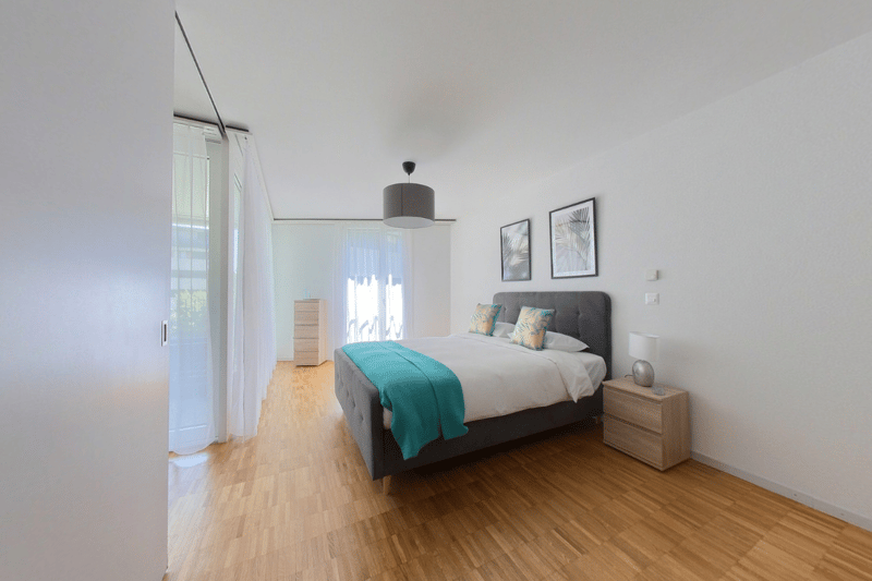 Bedroom with wooden floor, white walls, bed with pillows, nightstand with lamp, cabinet, and framed pictures on the wall.