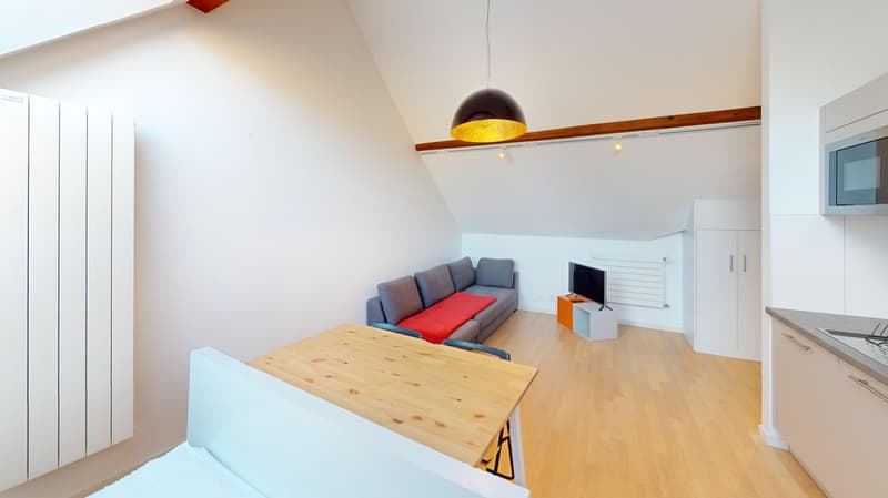 Bright and modern living room with vaulted ceiling, wooden beams, and a large window. The room features a gray sofa, a wooden dining table, and a TV stand. The lighting is provided by a pendant lamp with a golden interior.