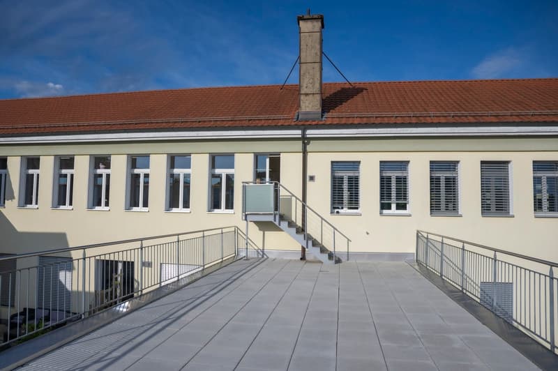 A two-story building with a red tile roof, beige exterior walls, and large windows. The building has a balcony or terrace with metal railings on the second floor. There is a set of stairs leading up to the entrance of the building.
