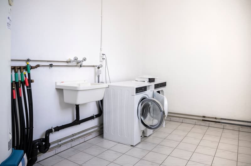 The image shows a laundry room with a washing machine, a utility sink, and various plumbing equipment and tools.