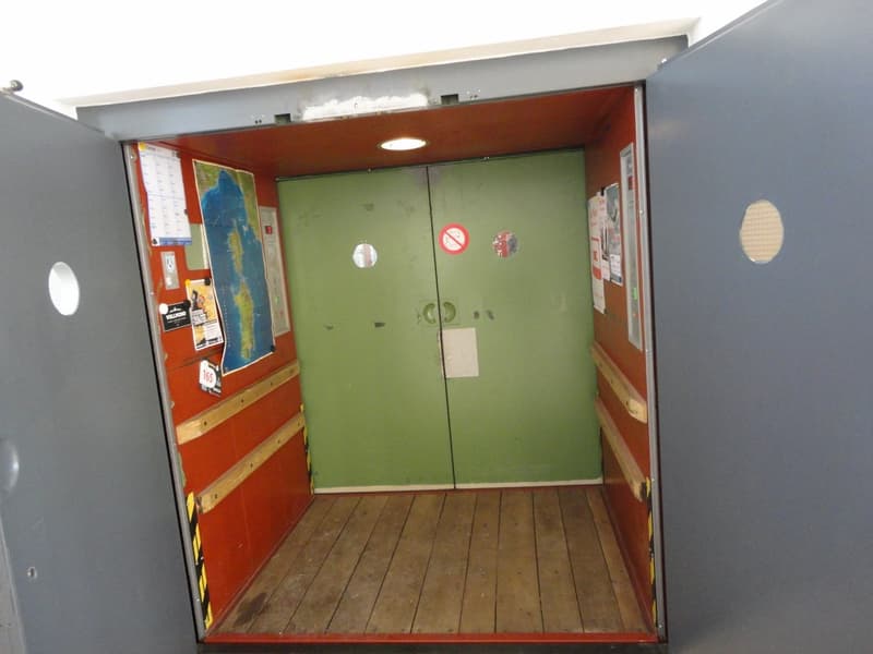 The image shows the interior of a room with a wooden floor and red and green walls. There are two green doors in the center of the room, and various items such as a map and posters are visible on the walls.
