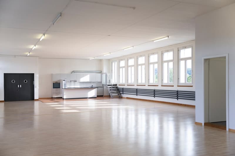 Large open space with hardwood floors, white walls, and multiple windows providing natural light. The room appears to be a multipurpose space with a kitchen area and a spacious open floor plan.