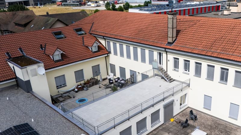 Multi-story apartment building with red tile roofs, white exterior walls, and balconies. There is a paved outdoor area with some furniture and a railing. The building appears to have parking spaces or garages on the ground level.