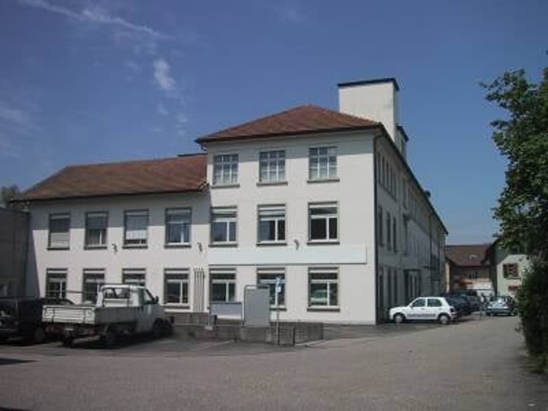 A multi-story commercial or office building with a white exterior and red tile roof. The building has several windows and appears to be located in an urban or suburban area with some trees and a parking area in front.