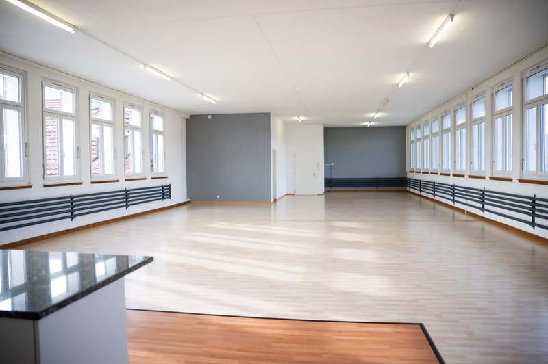 Large open space with hardwood floors, white walls, and multiple windows. The room appears to be a dance studio or similar type of space with wooden benches along the walls.