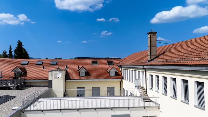 A multi-story building with a red tiled roof, dormer windows, and a balcony or terrace on the upper level. The building has a light-colored exterior and is surrounded by a clear blue sky with some clouds.