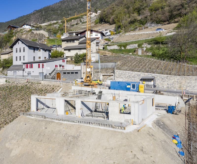 The image shows an active construction site in a mountainous, rural area. There is a large construction crane on the site, and several buildings and structures under construction. The surrounding landscape includes hills, trees, and other buildings in the
