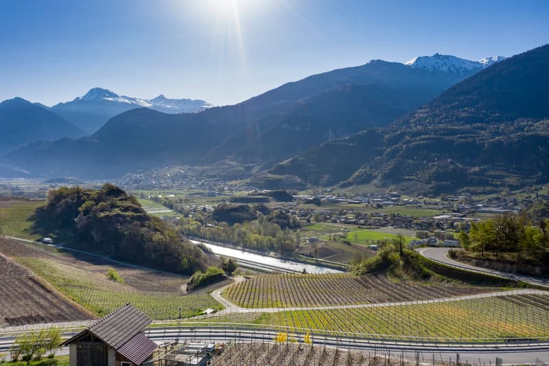 The image shows a picturesque mountain landscape with snow-capped peaks in the background. In the foreground, there are rolling hills covered in vineyards and farmland, with a small town nestled in the valley below. The scene is bathed in warm, golden sun
