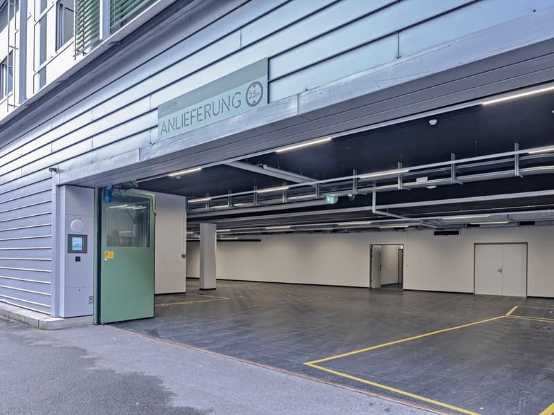Large indoor parking garage with concrete floors, yellow lines, and a sign indicating a 2.5m height limit. The garage has multiple entrances and exits, and appears to be well-lit with overhead lighting.