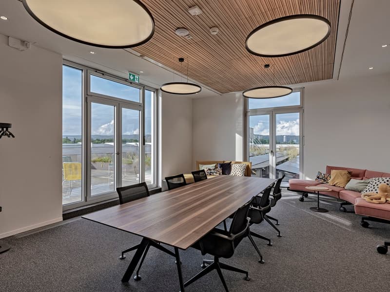 A conference room with a large wooden table, black chairs, wooden ceiling, large glass windows, white walls, and a couch.