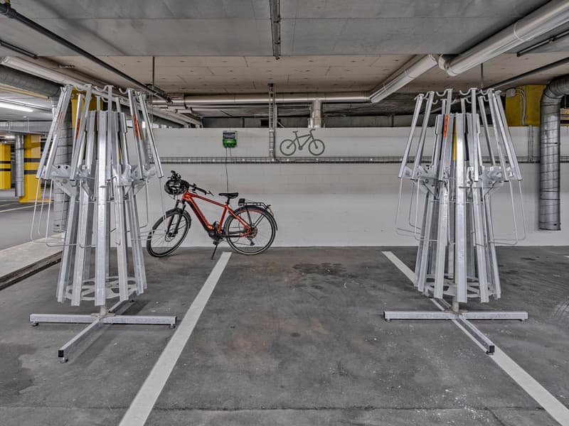The image shows a parking garage with multiple bicycle racks and a single bicycle parked in the center of the frame. The garage has a concrete floor and ceiling with exposed pipes and ductwork. The walls are plain concrete and there are yellow and black s