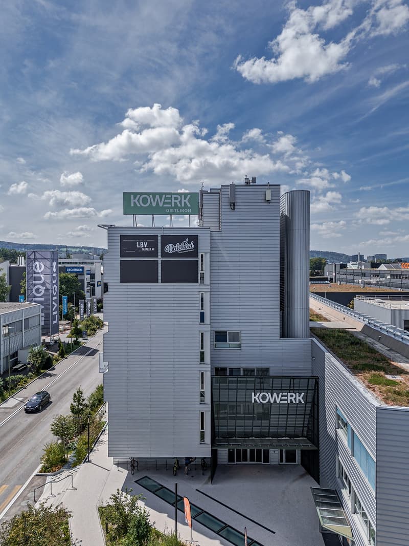The image shows a large, modern commercial building with the Kowerk logo prominently displayed. The building has a sleek, gray exterior with large windows and signage. There are other smaller buildings and structures visible in the surrounding area.