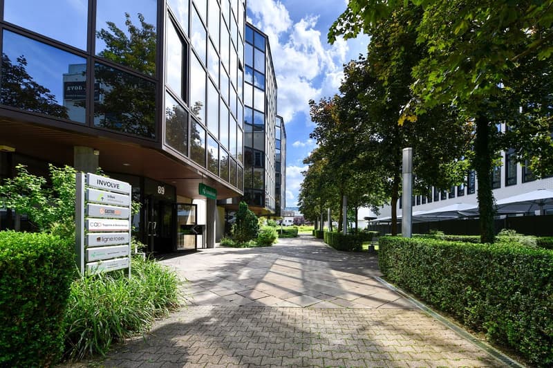 Multi-story office building with glass facade, surrounded by trees and greenery, with a paved walkway leading to the entrance.