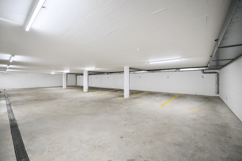 The image shows a large, open parking garage with concrete floors and white walls. The space is well-lit with fluorescent lighting, and there are several parking spaces marked with yellow lines on the floor. The garage appears to be clean and well-maintai