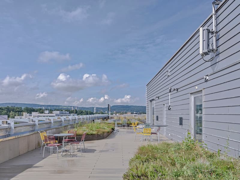 The image shows a multi-story commercial or industrial building with a rooftop terrace. The building has a gray siding exterior and the terrace features several colorful chairs and tables. The surrounding landscape includes grassy areas and a view of dist