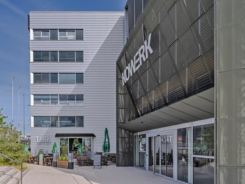 Multi-story commercial building with a large sign that says 'KONECRANES'. The building has glass windows and a patio area with tables and umbrellas in front. There is a parking area visible in front of the building.