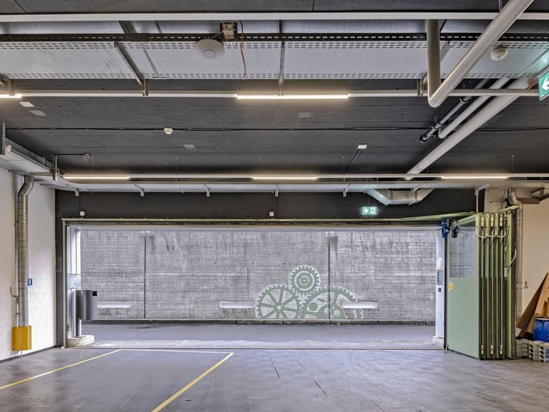 Large open warehouse space with concrete floors, exposed ceiling beams and ducts, and a large mural with gears and mechanical elements on the wall.