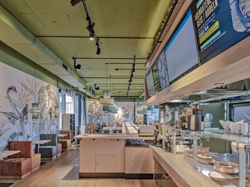 The image shows the interior of a cafe or restaurant. The space has a modern and stylish design, with wooden furniture, large windows, and a mural wall featuring tropical leaf patterns. There is a counter area with a display of food and drinks, as well as