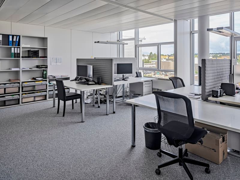 Spacious open-plan office with large windows providing a view, multiple workstations with computers and desks, shelving units for storage, and black office chairs.