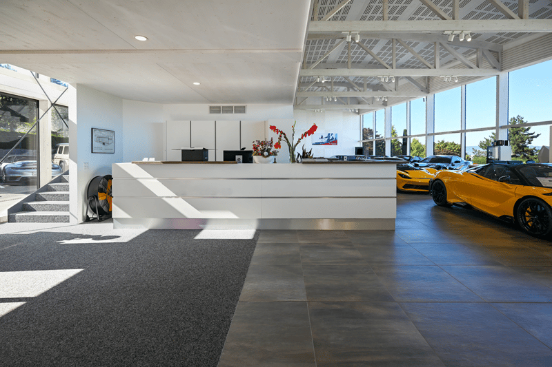 white walls, large glass windows, carpeted floor, desk with flowers, sports cars parked