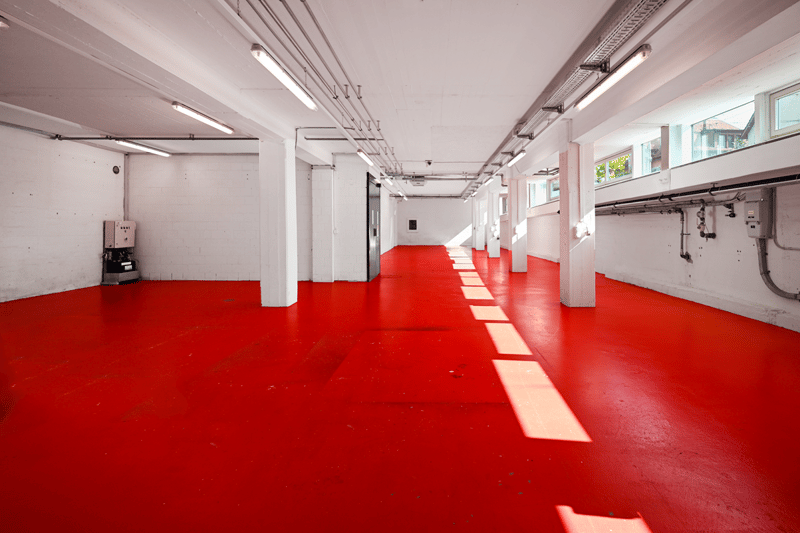 Red concrete floor, white walls, large windows, bright lighting, electrical box, white pillars