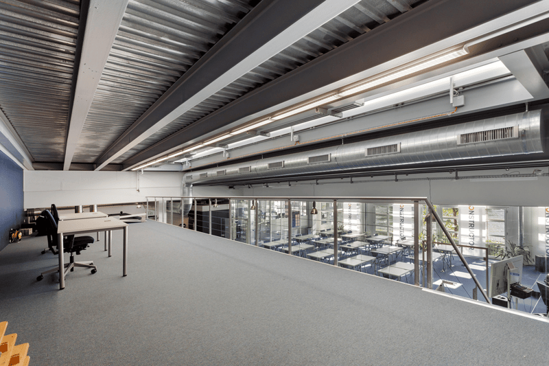 large empty room, several desks, gray flooring, white walls, glass partition, ceiling with ducts and lights