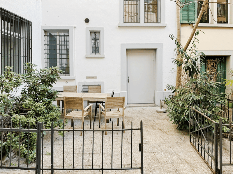 white building, wooden table and chairs, metal gate, plants, paved floor