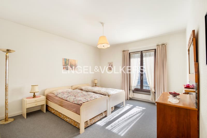 Spacious bedroom with large windows, white walls, hardwood floor, double bed, nightstands, dresser, and a pendant light fixture.