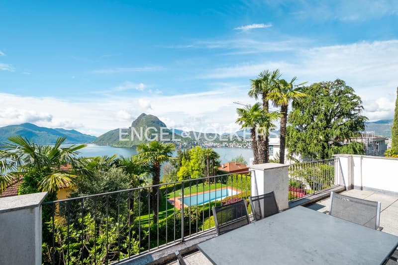 Luxury villa with panoramic view of Lake Lugano, surrounded by lush tropical vegetation including palm trees. The property features a swimming pool and a balcony or terrace overlooking the lake and the mountains in the background.