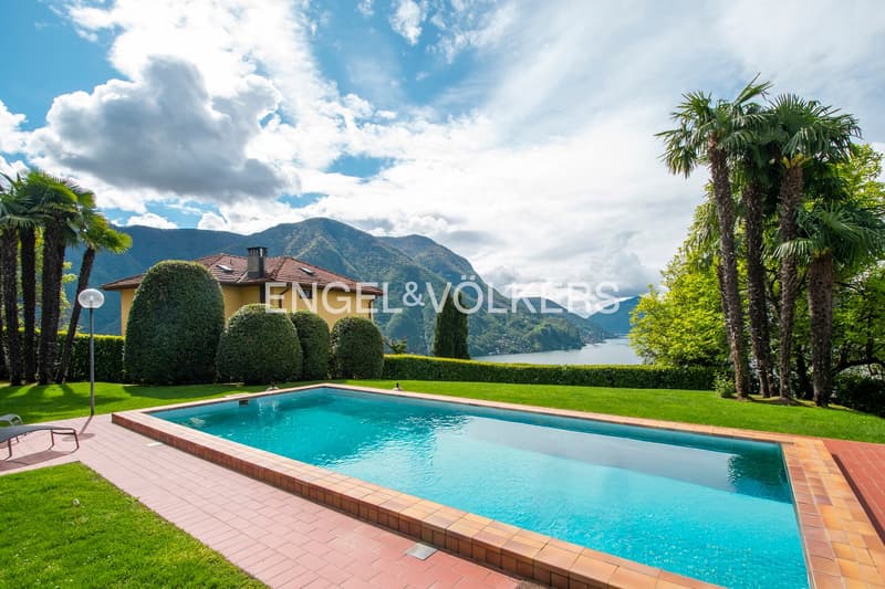 2-story house with red tile roof, surrounded by lush greenery and palm trees, with a large swimming pool in the foreground and a scenic lake and mountain view in the background