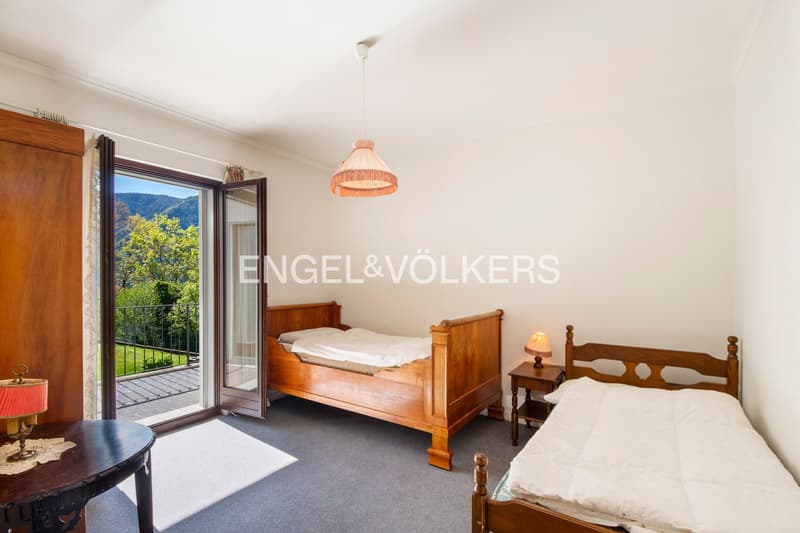 Spacious bedroom with wooden furniture, including a bed, nightstand, and dresser. Large windows provide a scenic view of the surrounding greenery. The room also features a pendant light fixture and a sliding glass door leading to a balcony or terrace.