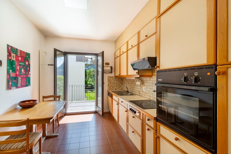 Spacious kitchen with wooden cabinets, tile flooring, black appliances including oven and stove, and a dining table. The kitchen opens up to a balcony or terrace with a view of the outdoors.
