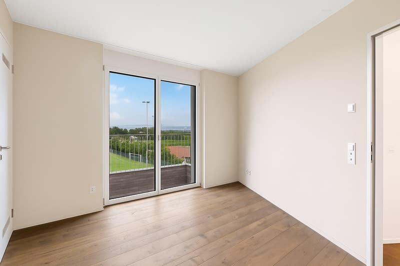 Wooden floor, large glass sliding doors, balcony with metal railing, neutral beige walls, electrical outlet on the wall, panoramic view of a grassy field with a tennis court